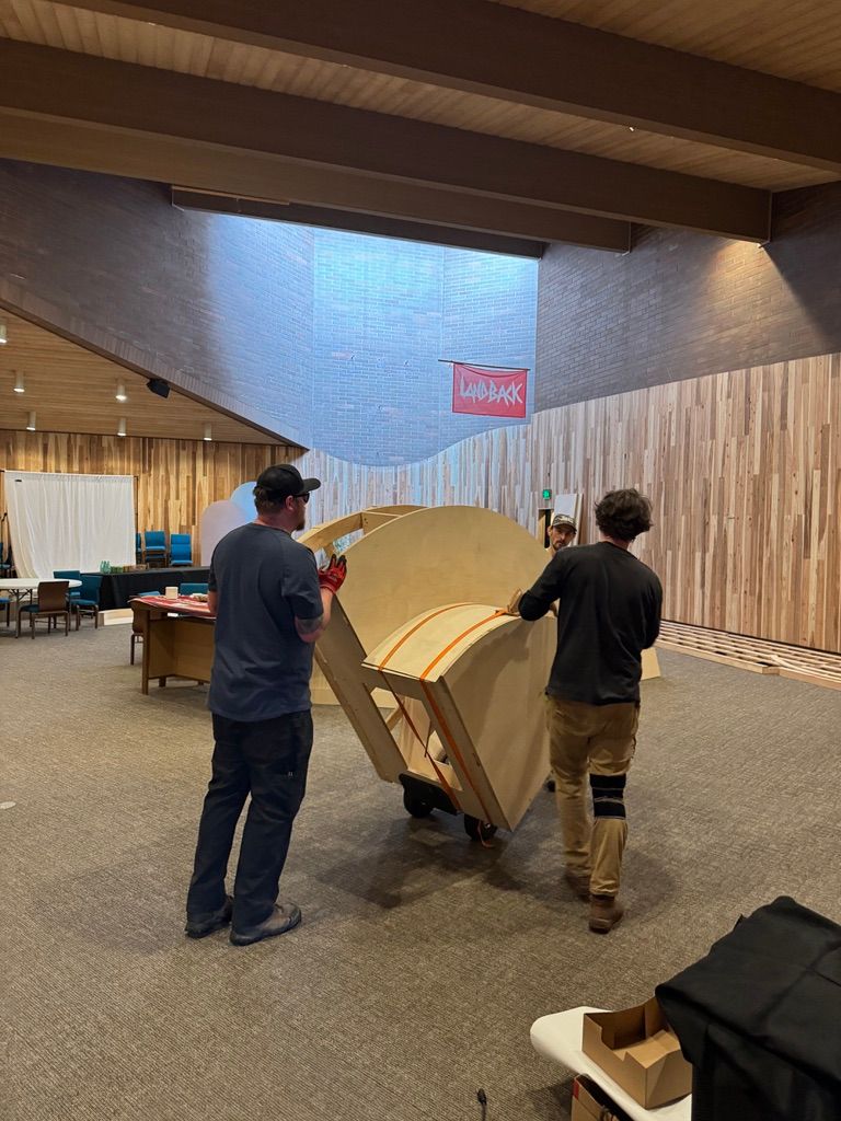 Two crew members wheel a curved wooden bench across the hall, under a red LANDBACK banner.