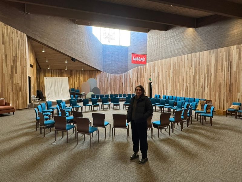 The renovated hall, its walls lined in new wood, with chairs set in a circle and a red LANDBACK banner high on the wall. One person stands in the middle.