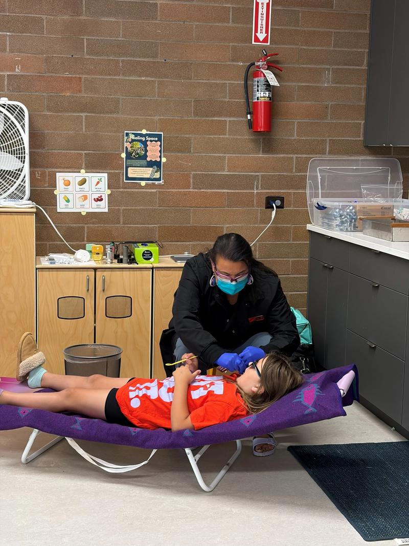 A dental hygienist in a mask checks a young child's teeth as the child reclines on a padded chair at a family wellness fair.