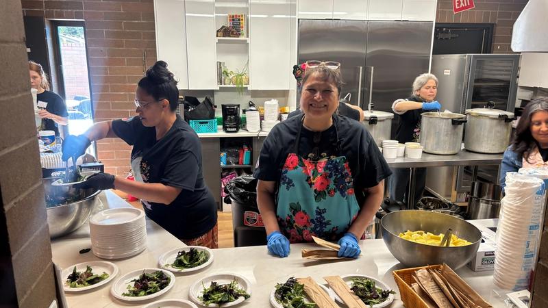 Two smiling volunteers in aprons and gloves plate traditional greens in a community kitchen.