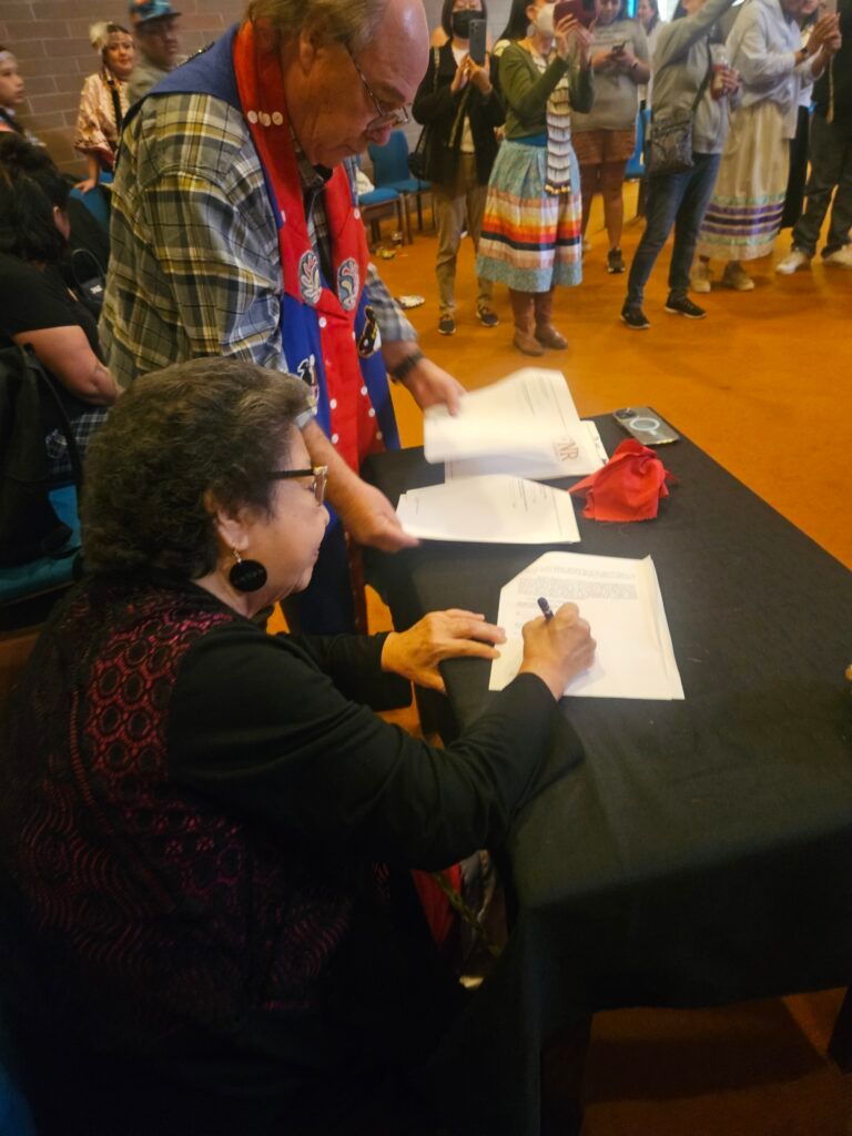 A woman signs the property transfer at a table while a man in a ribbon shirt stands beside her and community members in ribbon skirts look on.