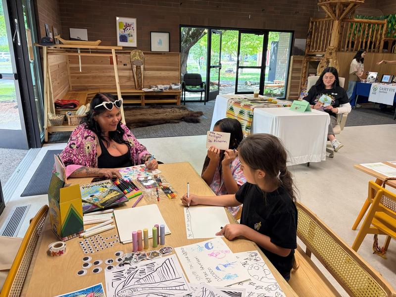 A caregiver and two young children sit at a craft table covered in coloring pages and markers at a family wellness fair.