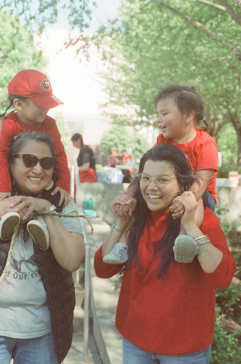 Two smiling women carry laughing toddlers in red shirts on their shoulders at a sunny outdoor community gathering.