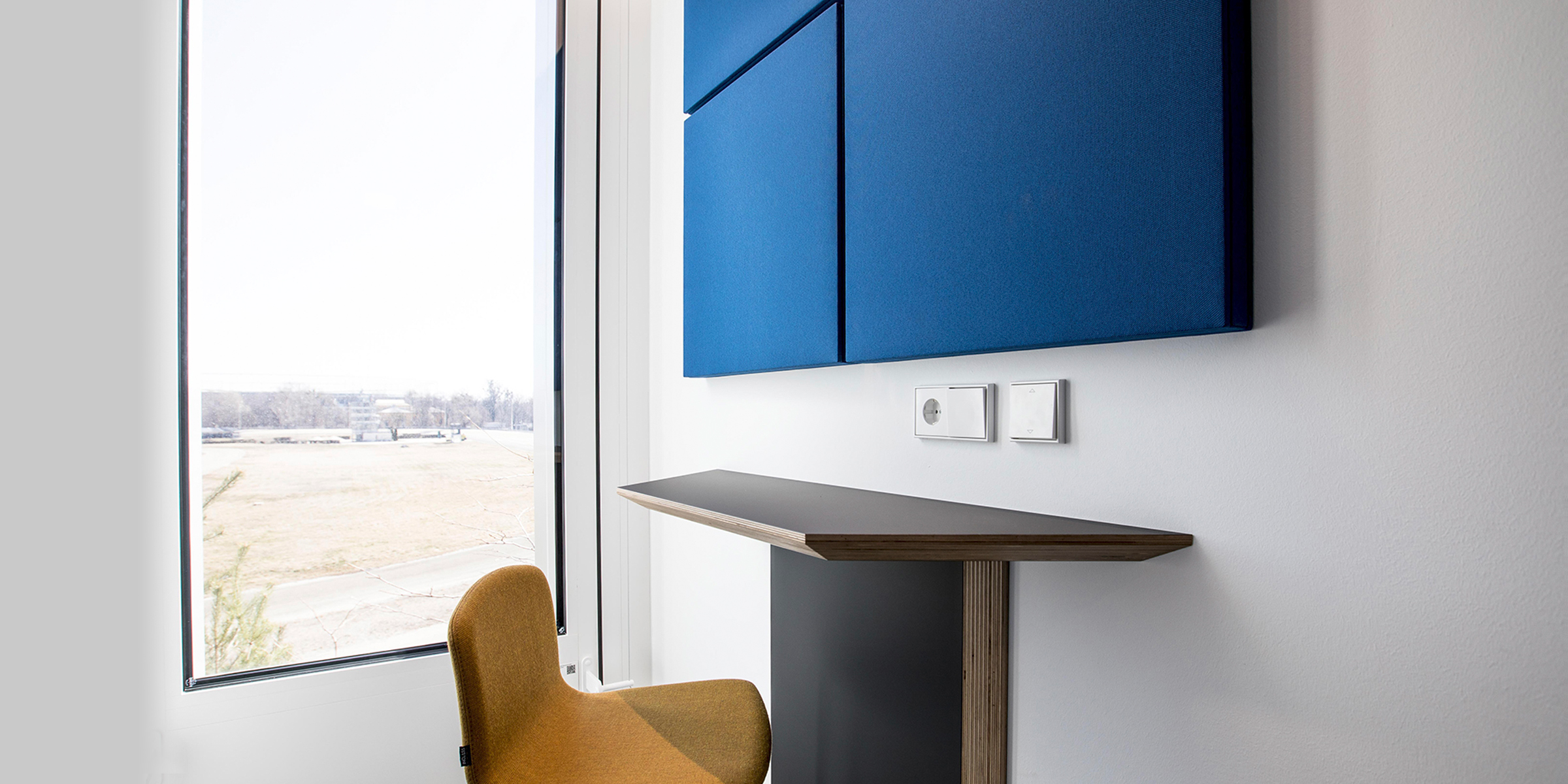 Ein minimalistisch eingerichtetes Büro mit einem großen Fenster, durch das man eine Aussicht auf eine flache Landschaft hat. An der Wand hängt ein großes, blaues Schallabsorptionspaneel und darunter befindet sich ein schmaler Schreibtisch mit einer braunen Sitzschale. An der Wand ist ein weißer Lichtschalter zu sehen.