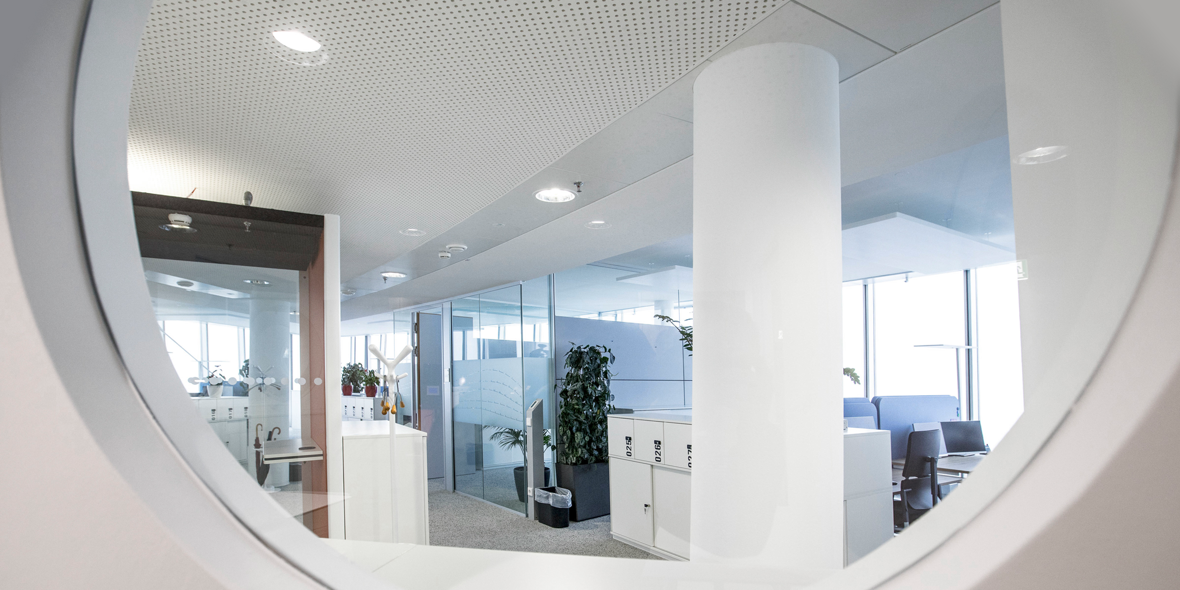 Blick durch ein rundes Bullauge auf ein helles Bürointerieur mit weißen Säulen, Büroschränken und Arbeitsbereichen im Hintergrund.