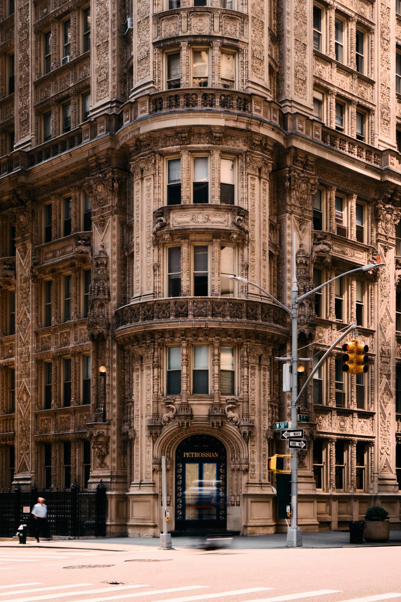 A large, ornate building with multiple levels and balconies stands on a city street corner, with a traffic light and pedestrian crossing nearby.
