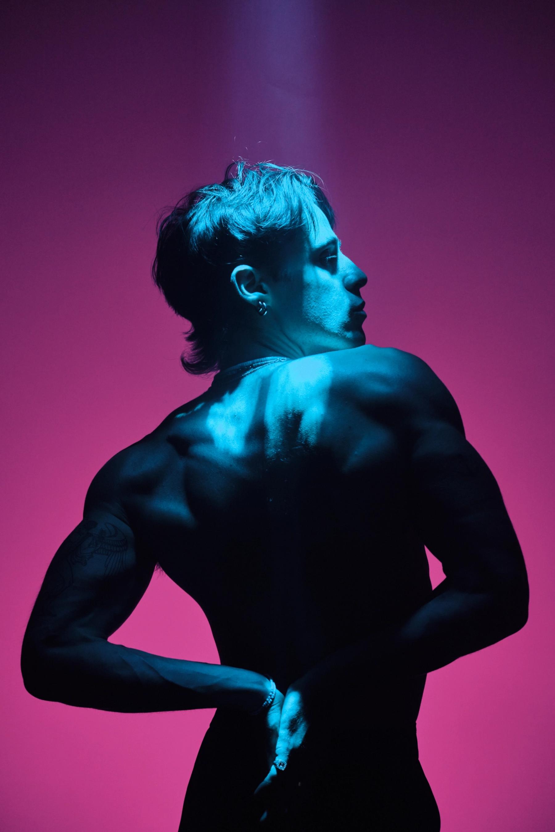A man with a black shirt and dark hair stands confidently in a pinkish-red background, his hands on his hips and arms outstretched.