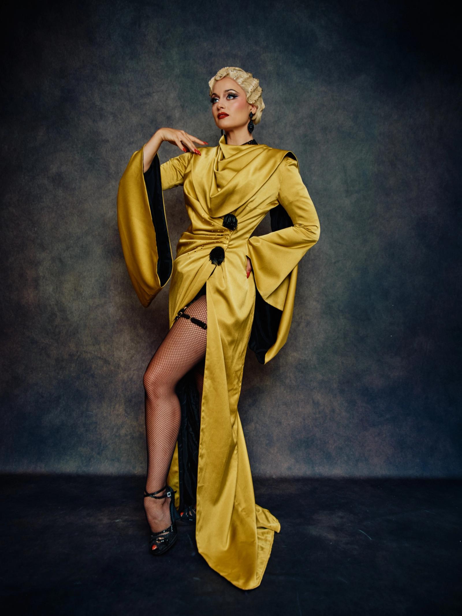A woman in a golden dress with a black belt and a red hairdo stands confidently against a dark blue background.