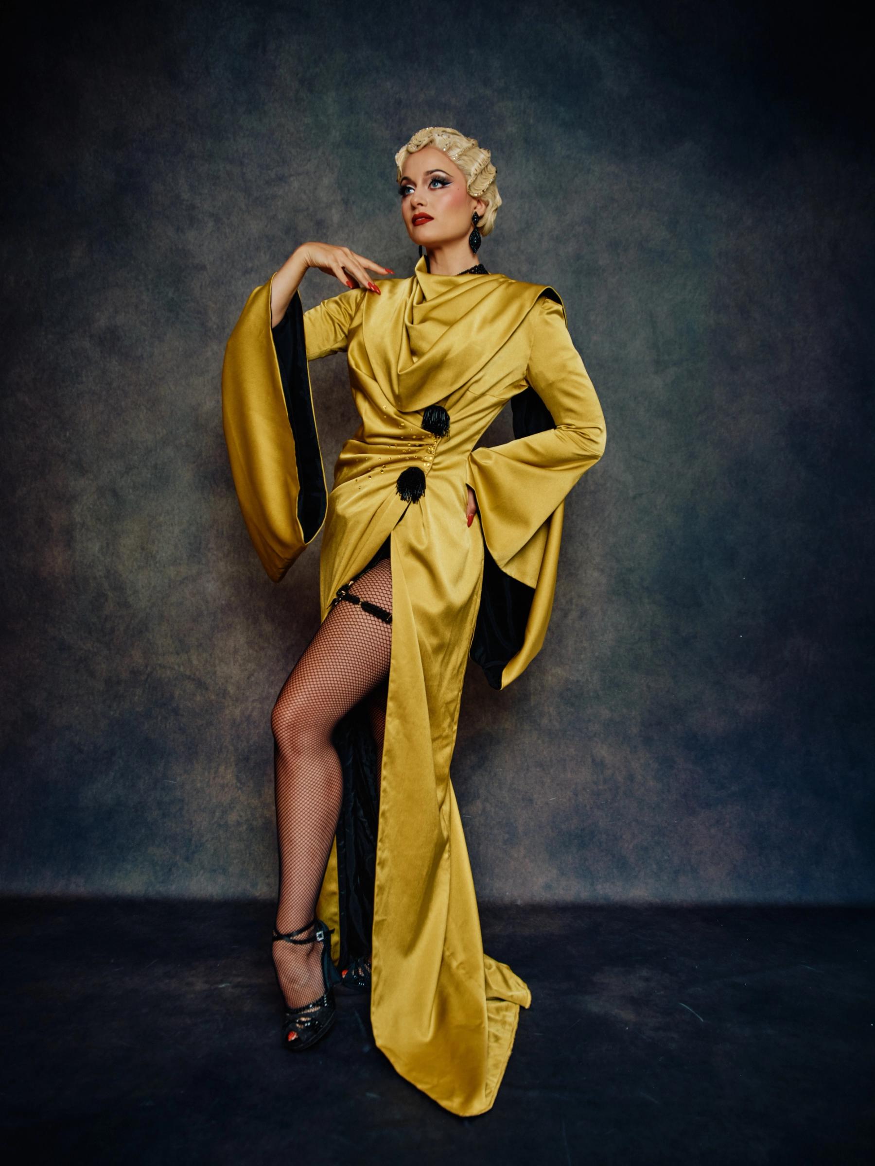 A woman in a golden dress with a black belt and a red hairdo stands confidently against a dark blue background.