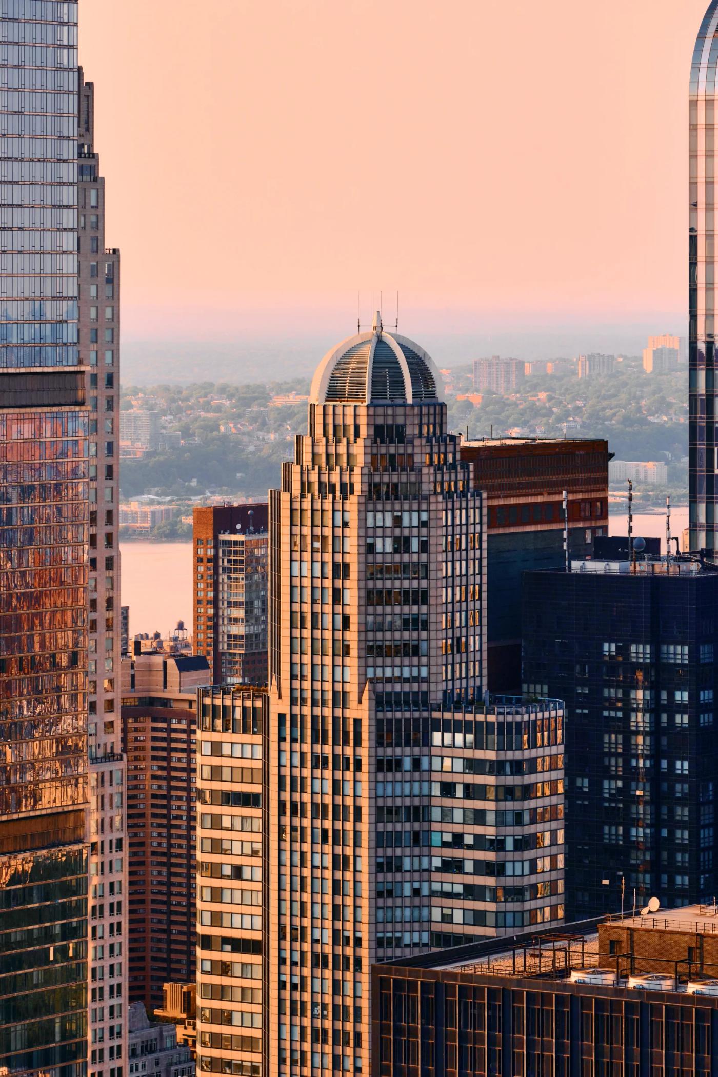 A cityscape at sunset features a mix of modern and older buildings, with a prominent dome-shaped building in the foreground. The sky is a warm pink, a