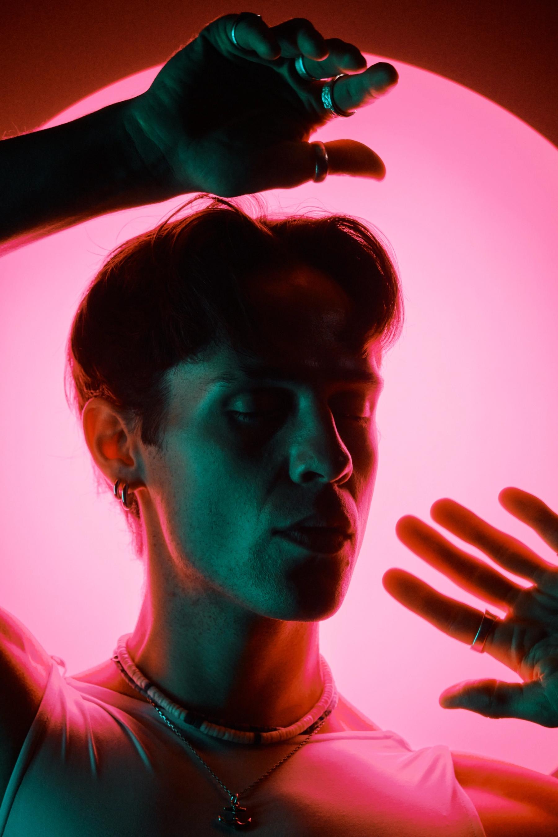 A young man in a white shirt and necklace stands in front of a pink neon background, reaching out with one hand while his other hand rests on his hip.
