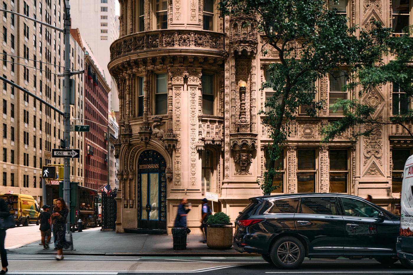A bustling city street features a white ornate building with intricate carvings, a parked black SUV, and pedestrians walking on the sidewalk.