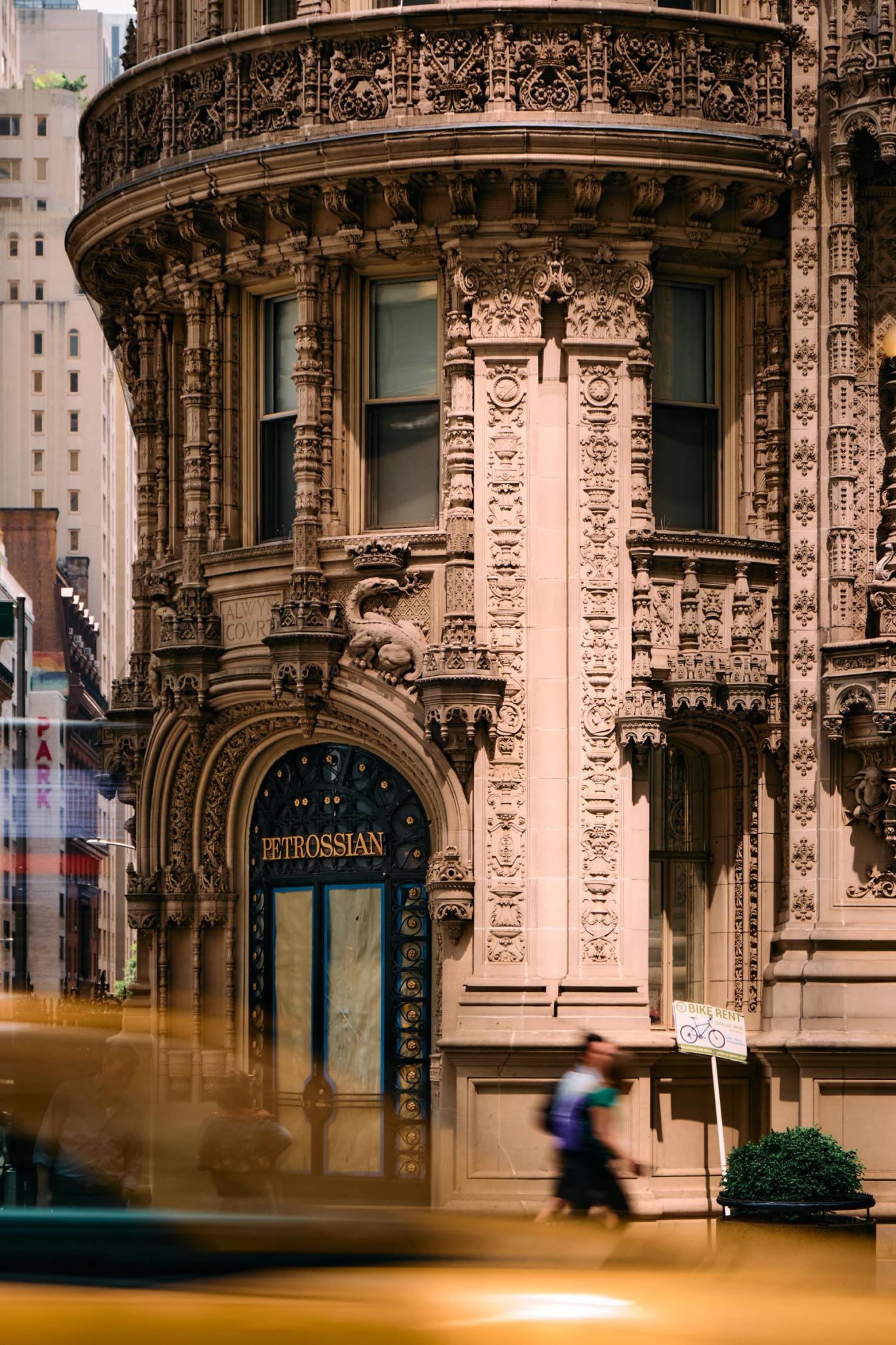 A bustling city street scene features a man crossing the street, a parked yellow taxi, a tall, ornate building, and a traffic light.