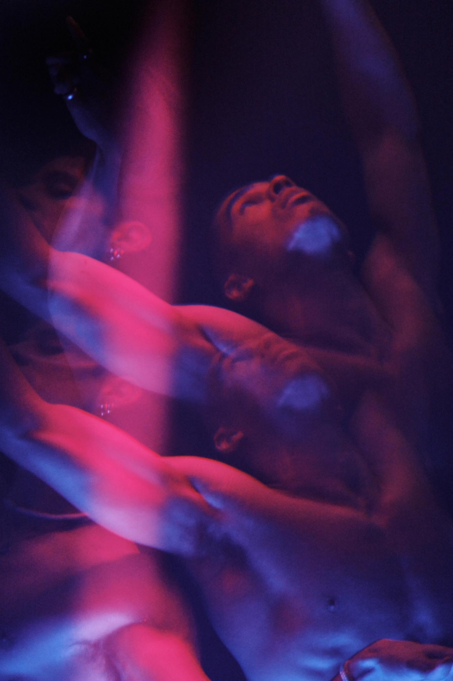 A man is shirtless, holding a microphone, and looking upwards. A woman is in the background, also shirtless.