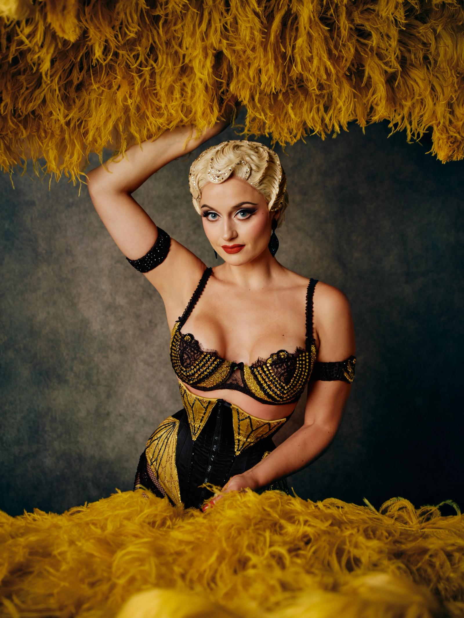 A woman with blonde hair, wearing a black corset with gold accents and a black lace-up top, poses elegantly with her hand on her head and her hair sty