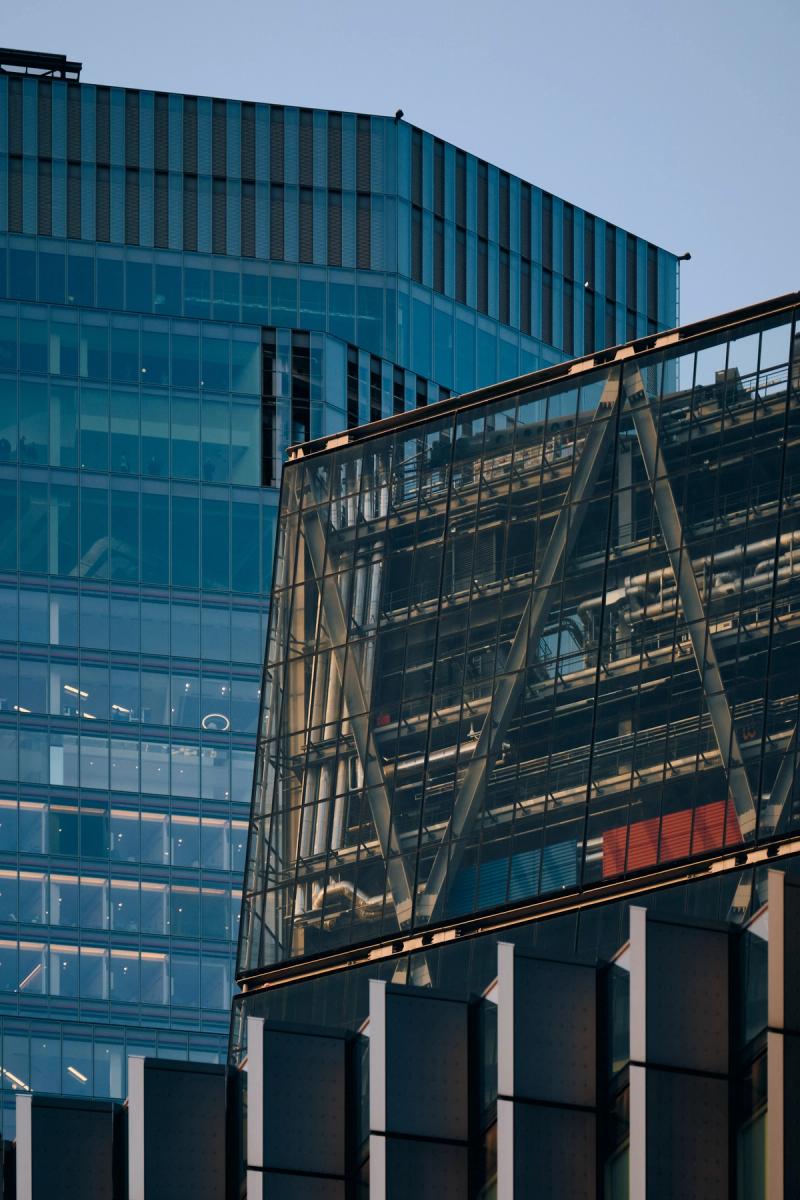 Modern glass office buildings with teal facade and industrial steel-framed connector bridge with diagonal cross-bracing, City of London
