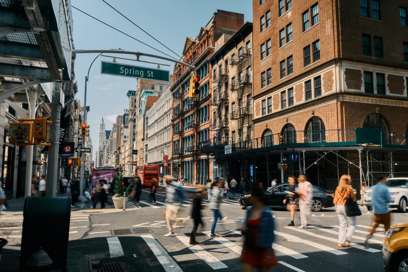 A bustling New York City street corner features brick buildings, pedestrians, and cars, with a clear blue sky.