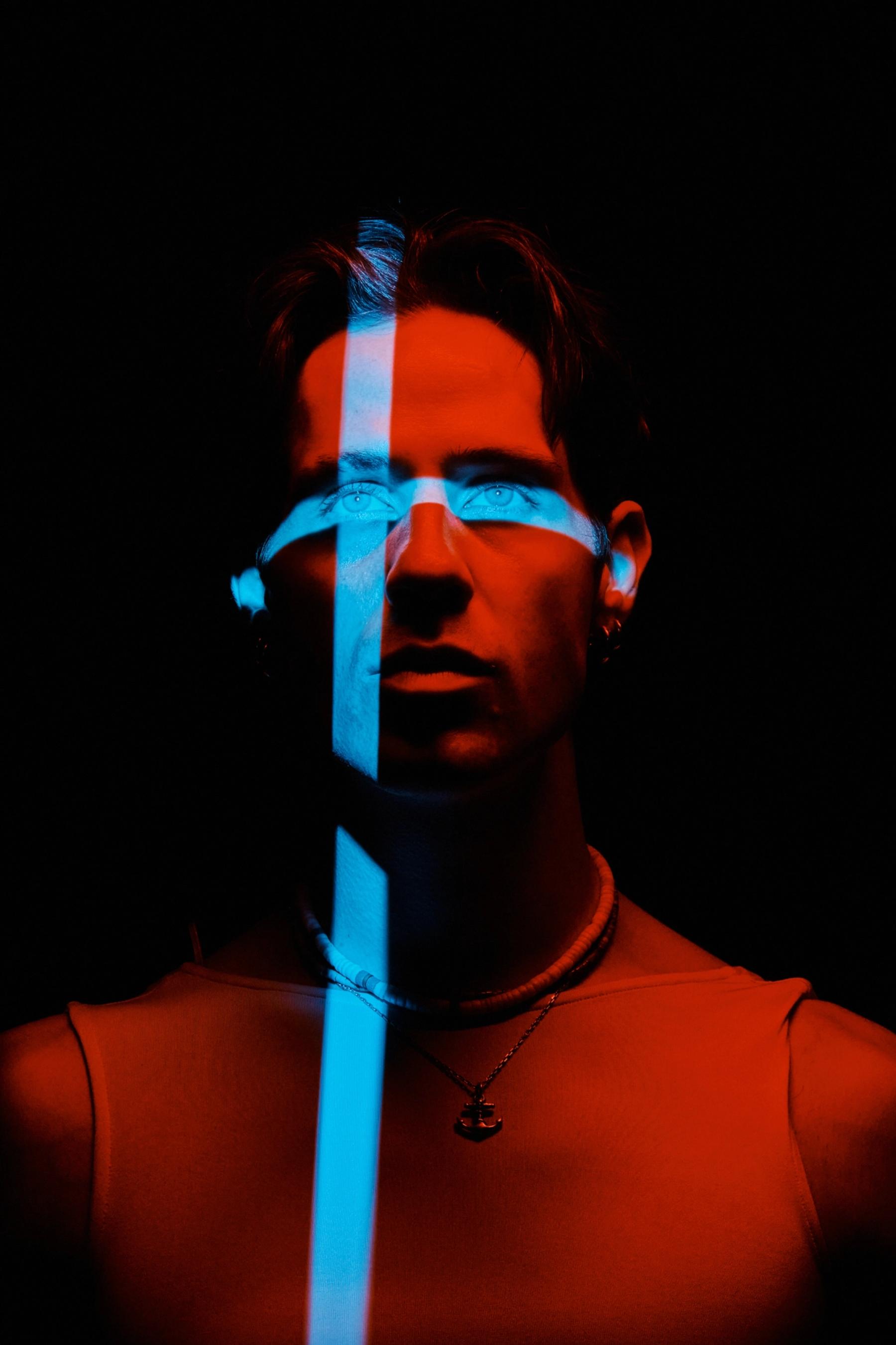 A young man with a blue light on his head and a necklace stands in a dark room, gazing directly at the camera.