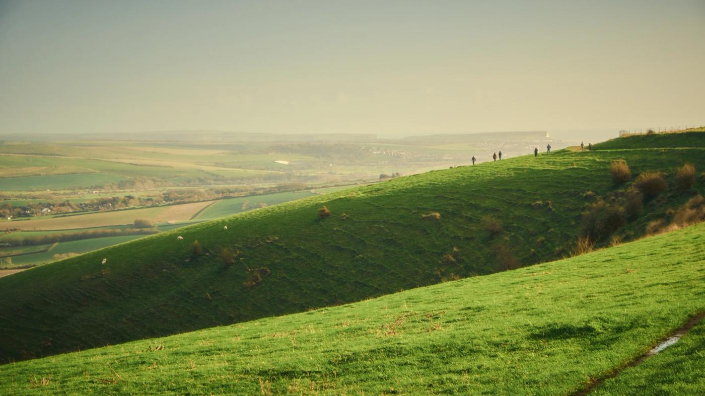 A serene countryside scene features a lush green hill with a few people, a valley with rolling hills, and a hazy blue sky.