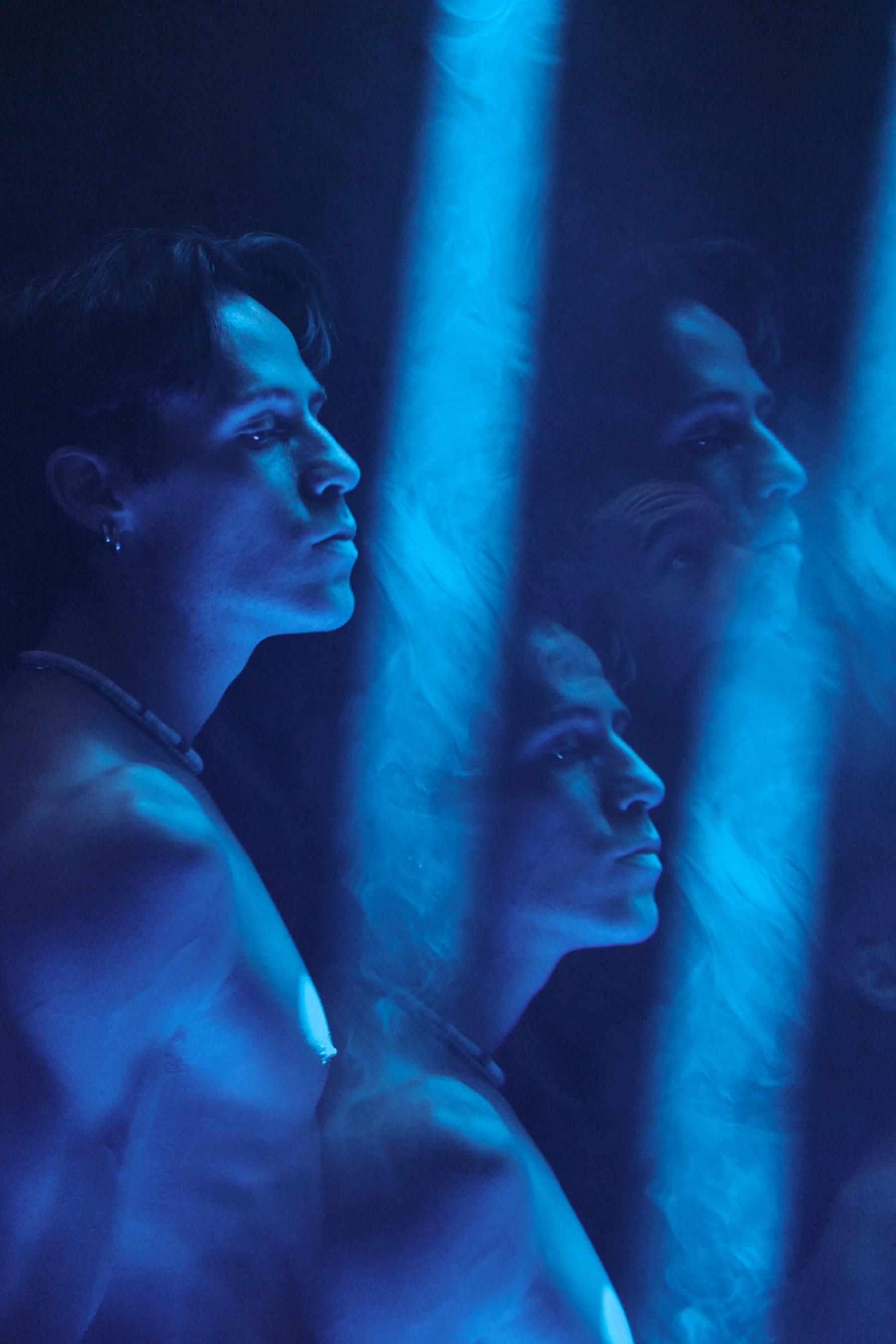 A woman with a necklace and earrings, looking at her reflection in a mirror, is in a dimly lit room with blue lights.