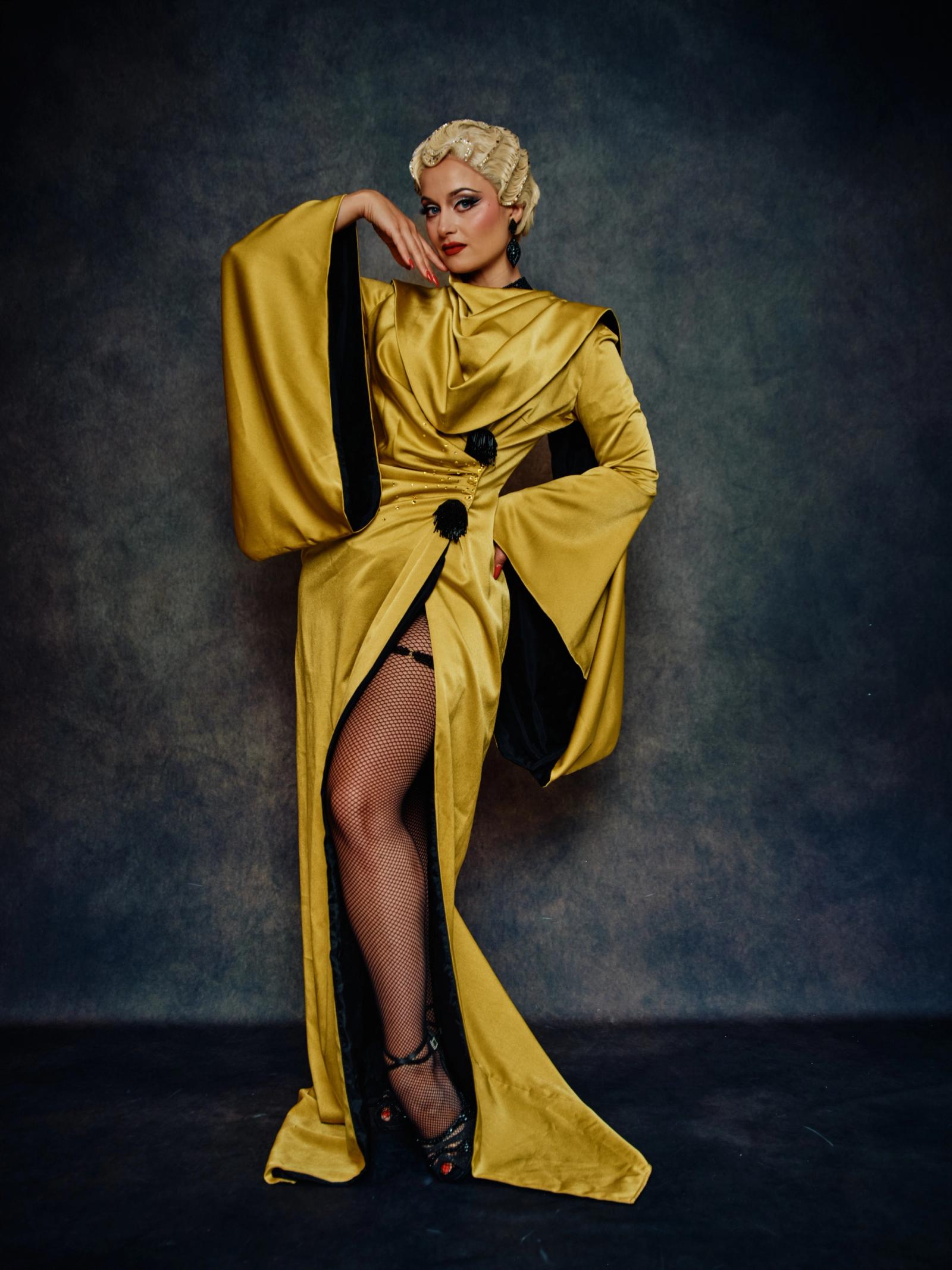 A woman in a golden, flowing dress with a black bow and black high heels stands confidently against a dark blue background.