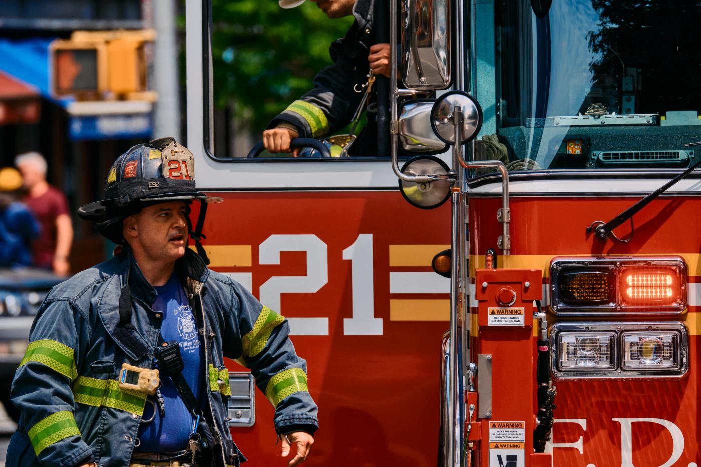 A firefighter in a blue and yellow striped jacket and helmet stands in front of a red fire truck with the number "100" on the side. The truck has a la