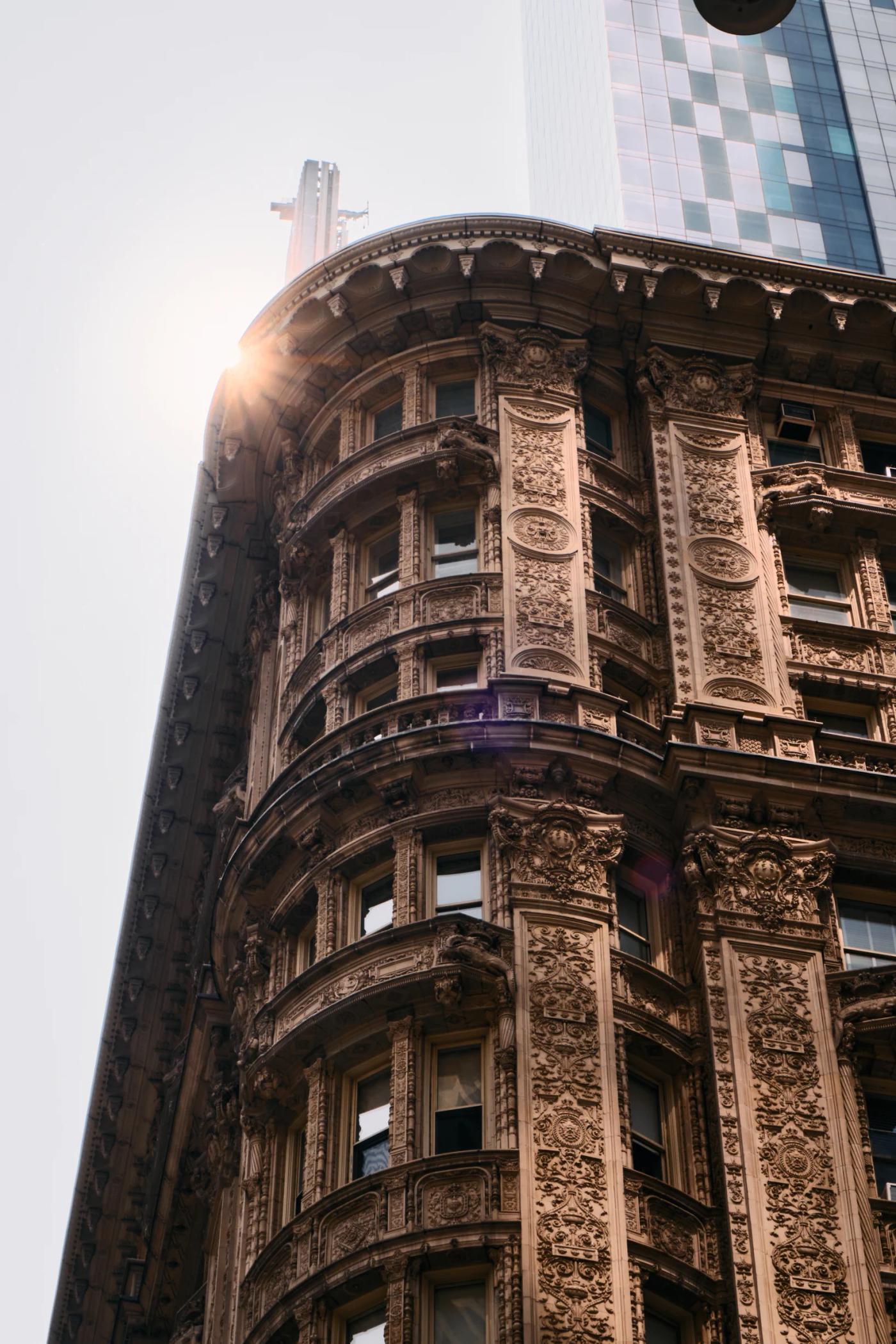 A tall, ornate building with intricate carvings and a clock on its facade stands in a bustling cityscape, with a tall skyscraper in the background.