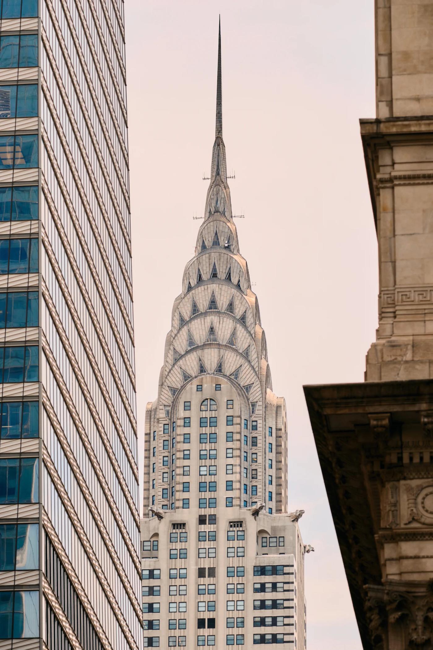 The Empire State Building, a white skyscraper with a pointed spire, stands tall in a bustling cityscape, surrounded by other buildings and a pale blue