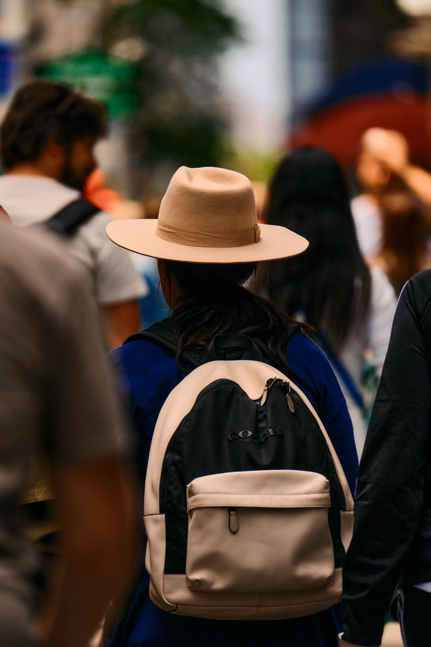 A woman in a beige hat and backpack walks down a busy street, surrounded by other people.