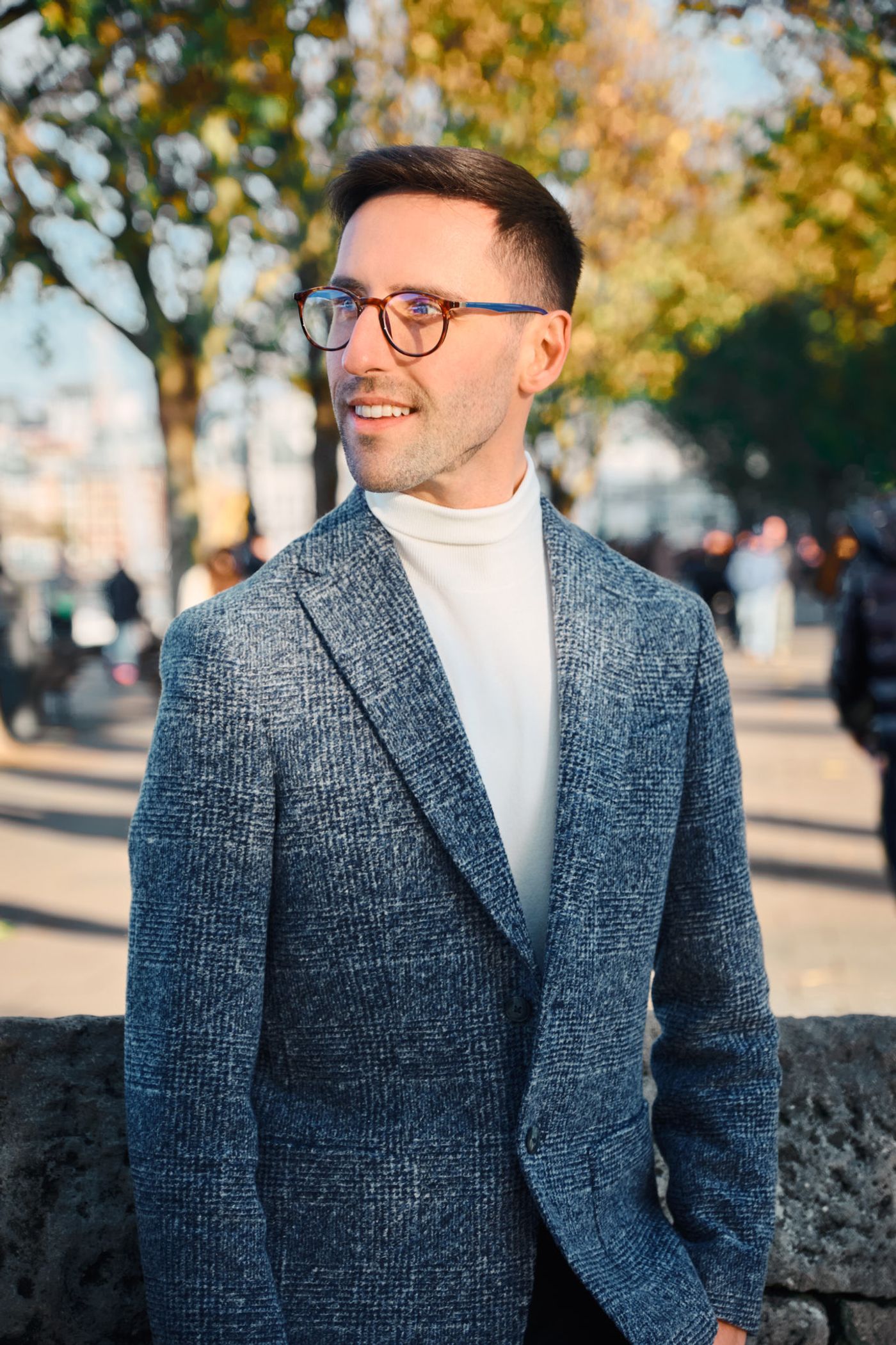 A man in a gray blazer and white turtleneck stands on a sidewalk, smiling and looking to the left.