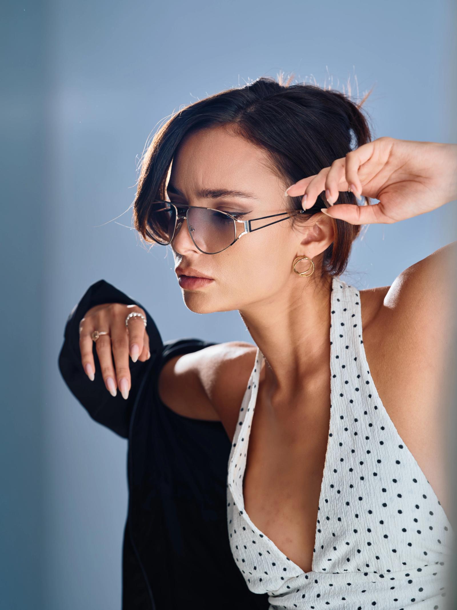 A stylish young woman in a polka dot top and sunglasses poses for a photo against a blue background.