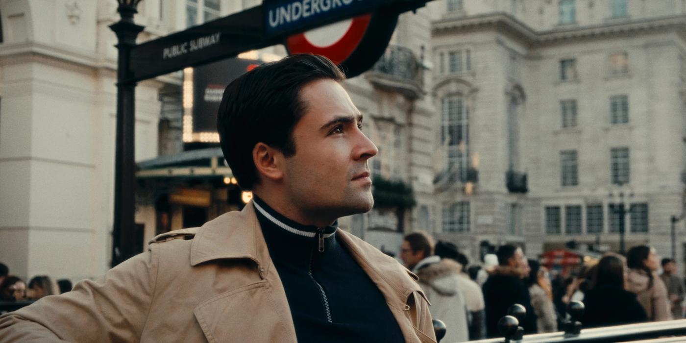 A man in a beige trench coat and black shirt, with short dark hair, stands in front of a London Underground sign, gazing upwards.