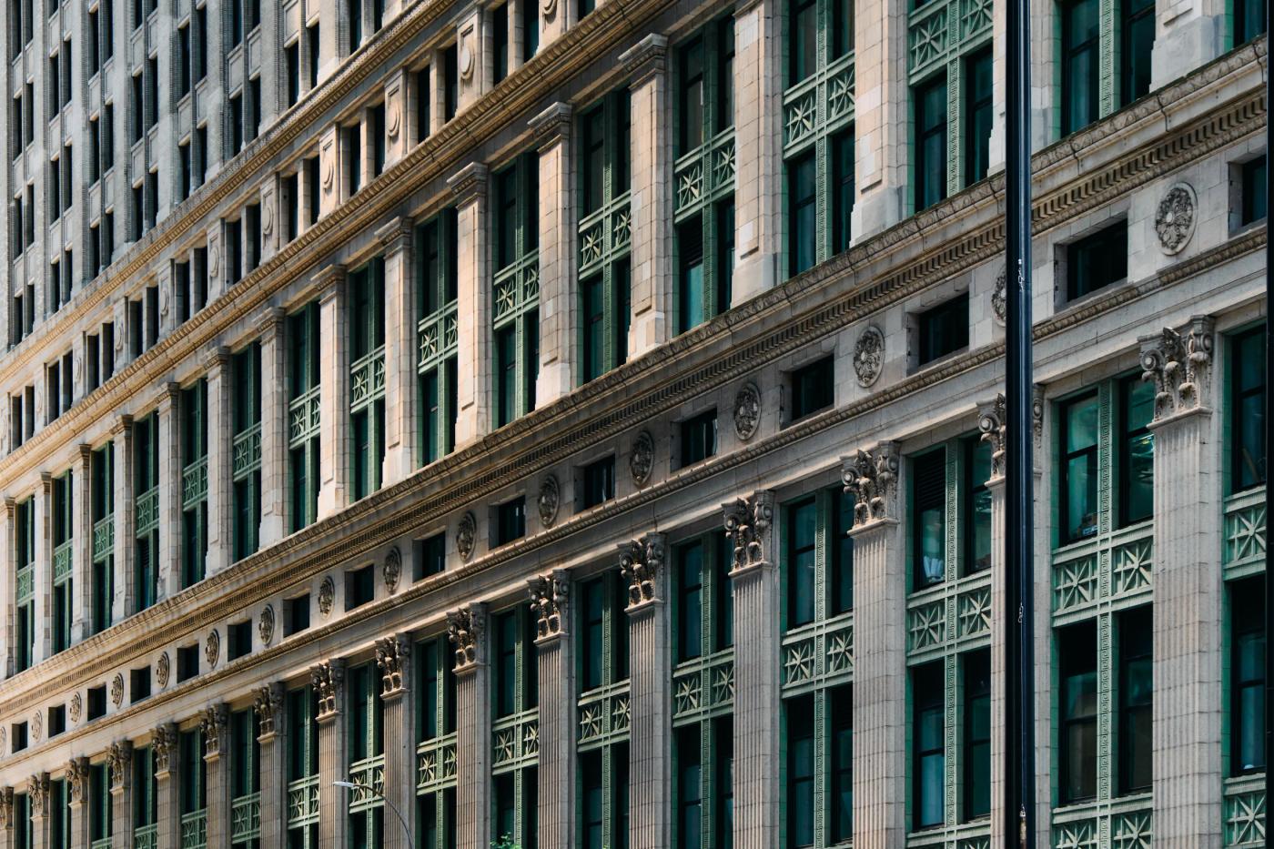 A tall, white building with green columns and a curved facade stands in a bustling city, surrounded by other buildings and a street lamp.