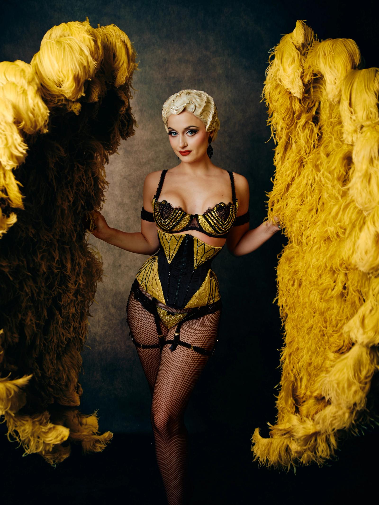 A woman in a black and yellow corset, with a white headband and blonde hair, poses elegantly in front of two large, yellow feathered objects.