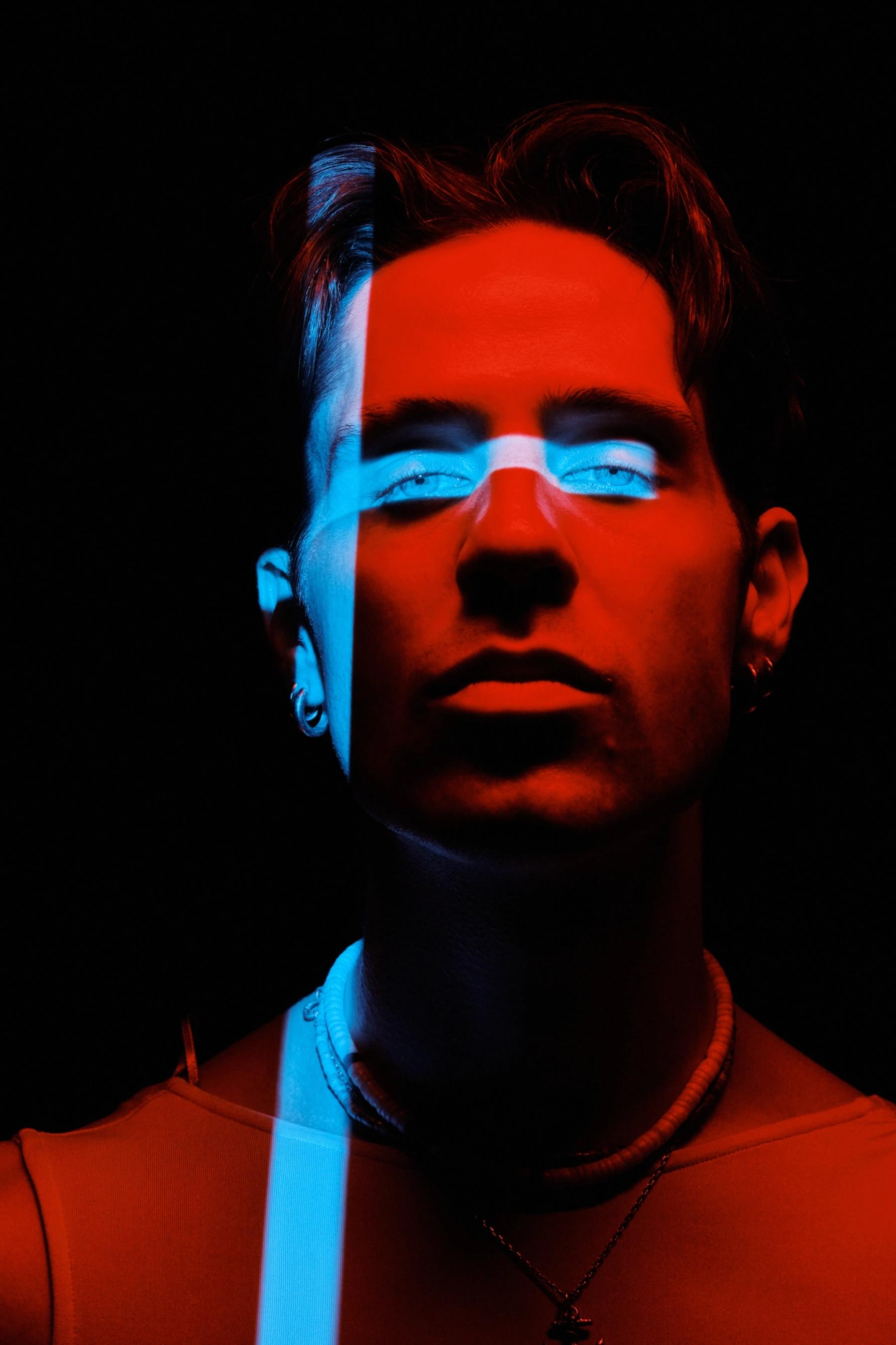 A young man with glasses and a necklace is in a dark room, looking directly at the camera. The background is black, and the man's face is illuminated 