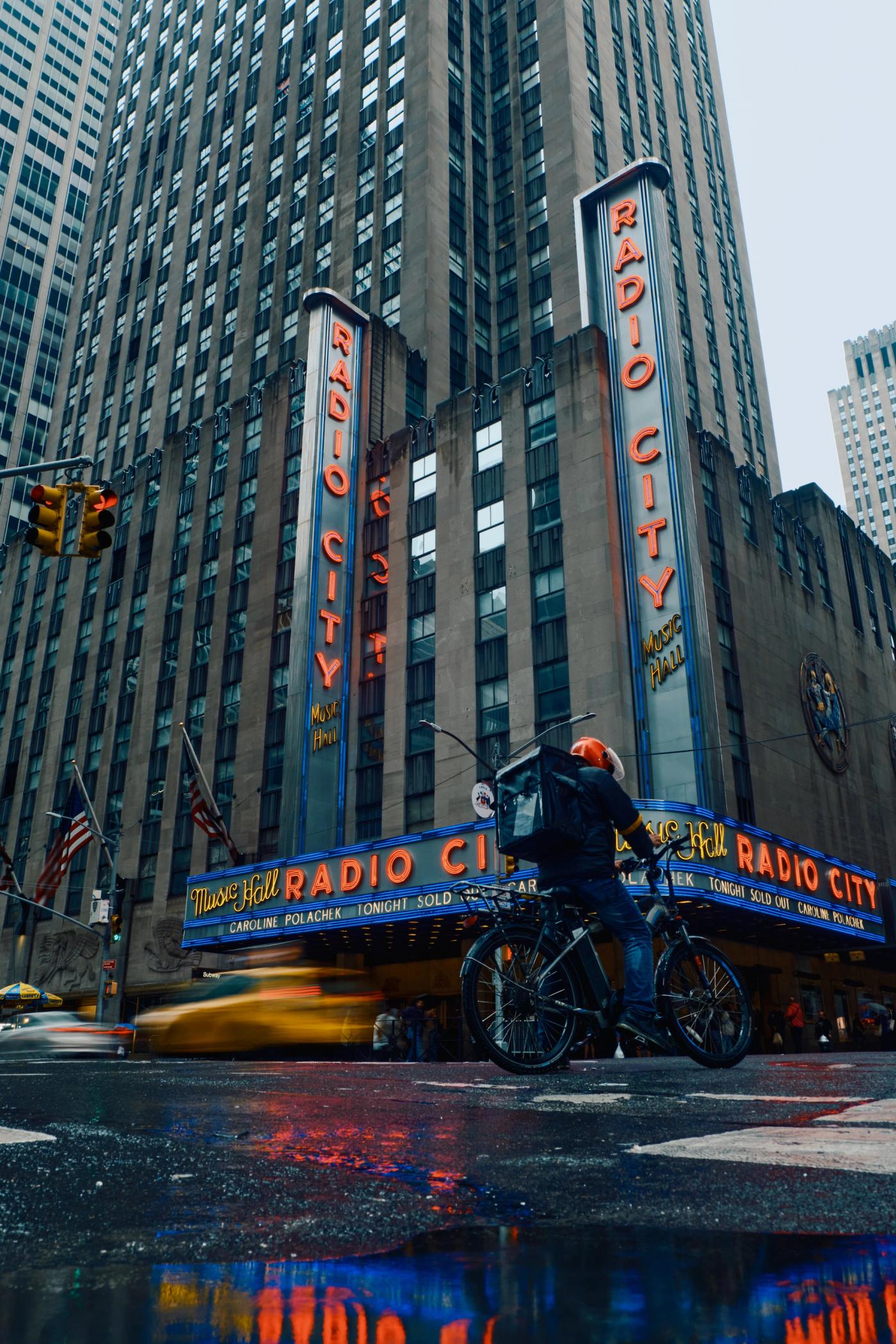 The image captures a bustling New York City street scene, featuring the iconic Radio City in New York City. The building, known as "MAX 100," is adorn
