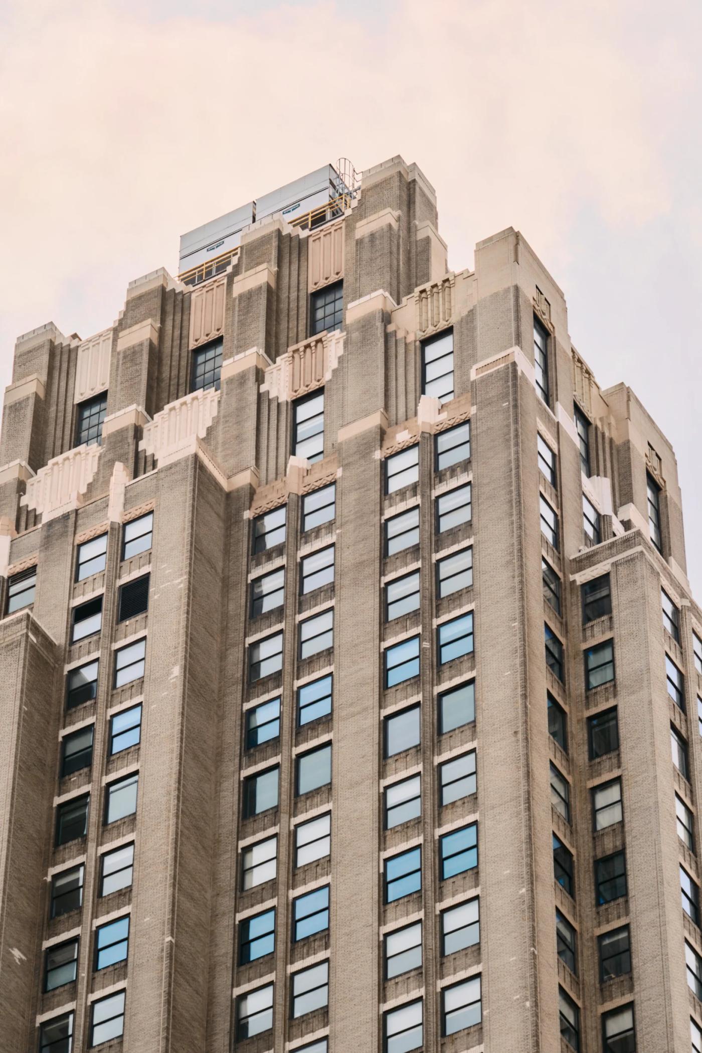 A tall, beige building with numerous windows stands against a cloudy sky, featuring a clock on the right side.