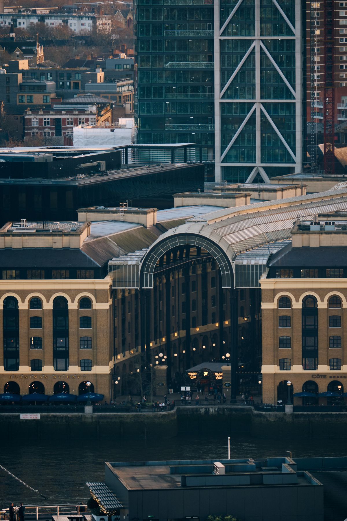 Aerial view of historic ornamental copper spire above modern waterfront promenade with contemporary office buildings, London golden hour
