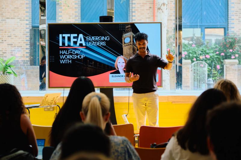 A man stands in front of a TV screen, talking to a group of people.