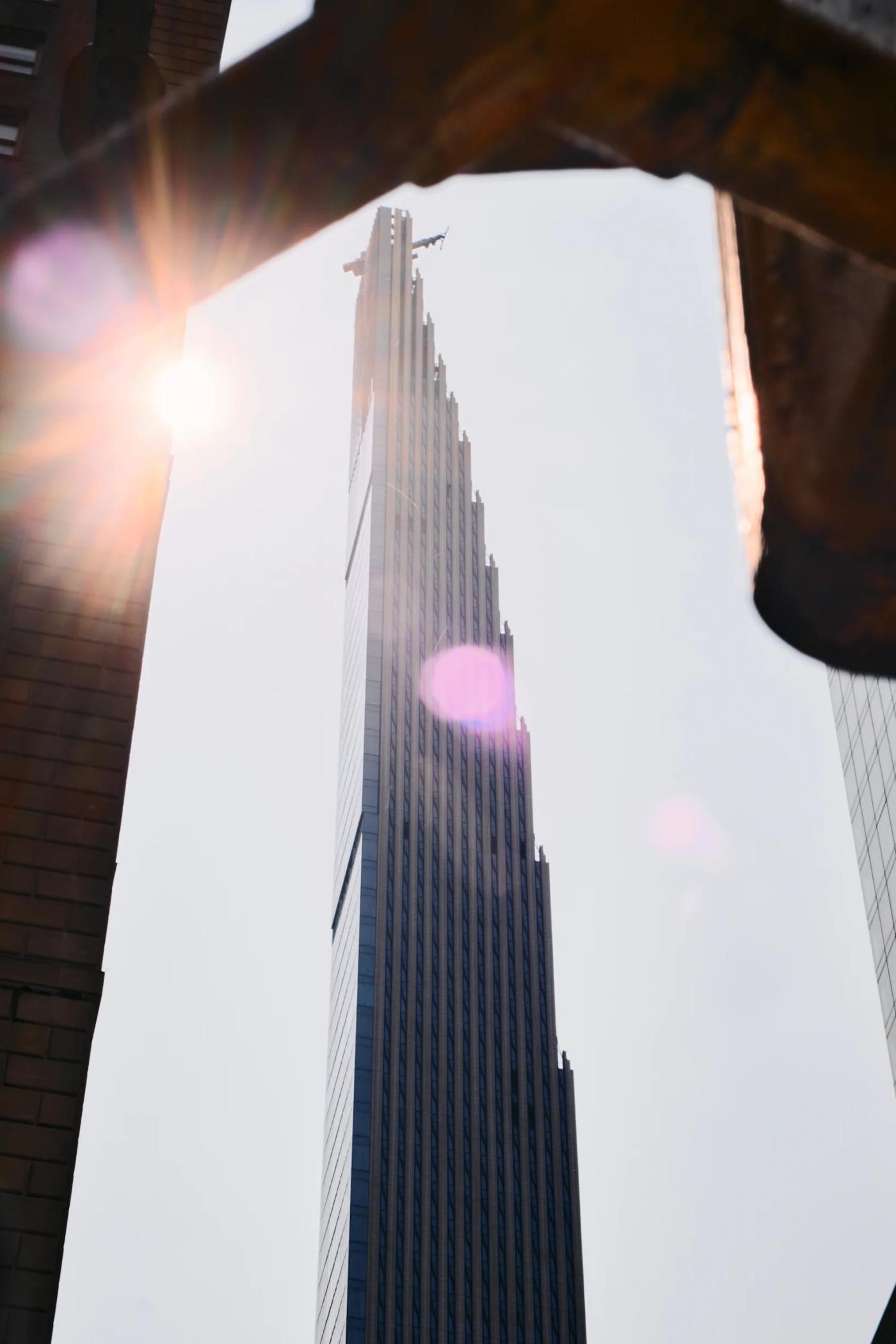 A tall, modern skyscraper with a pointed top and vertical lines dominates the cityscape, surrounded by other buildings and a sunlit sky.