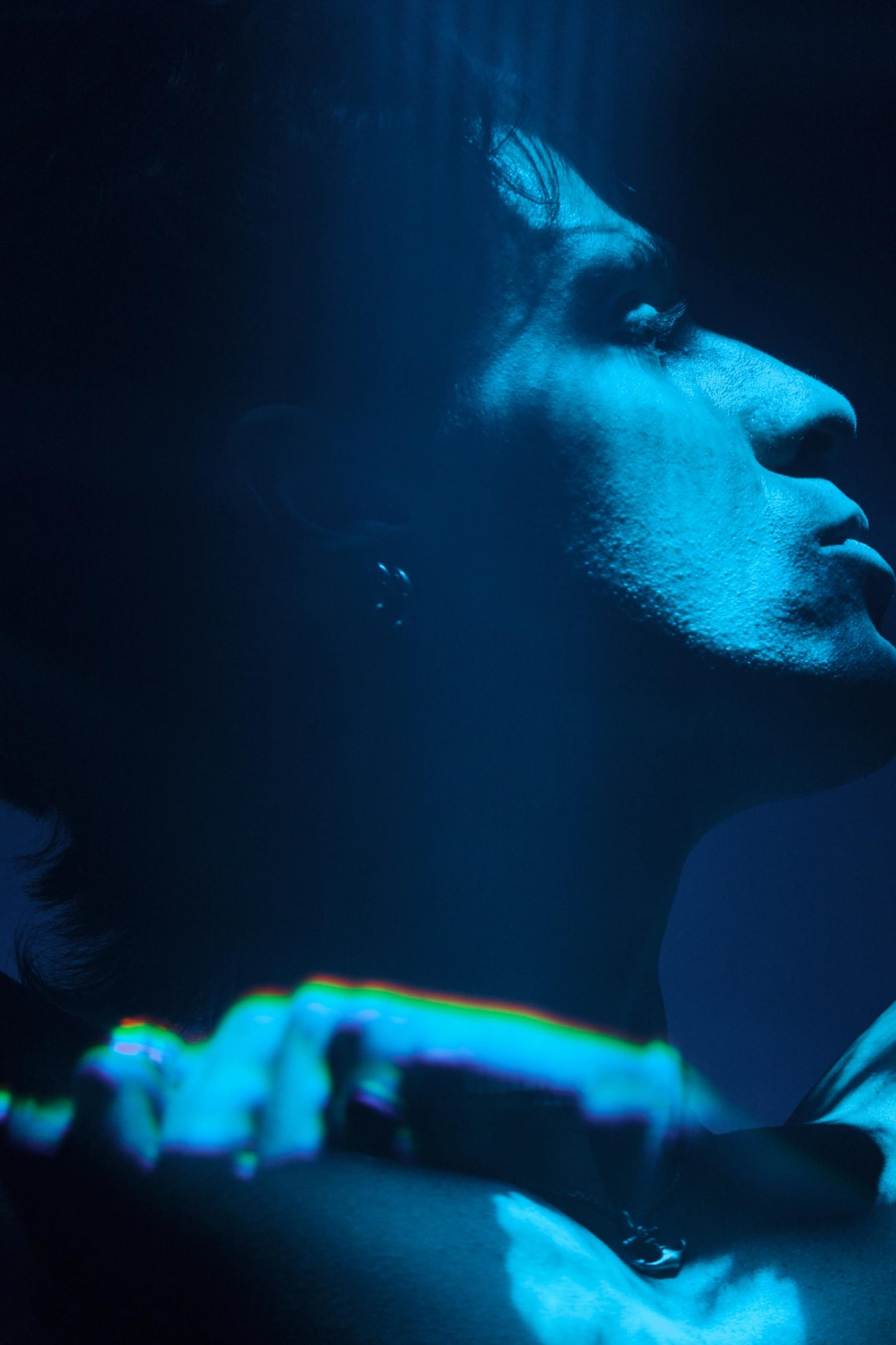 A man with dark hair and a necklace is in a dark blue room, gazing upwards with a mysterious expression.