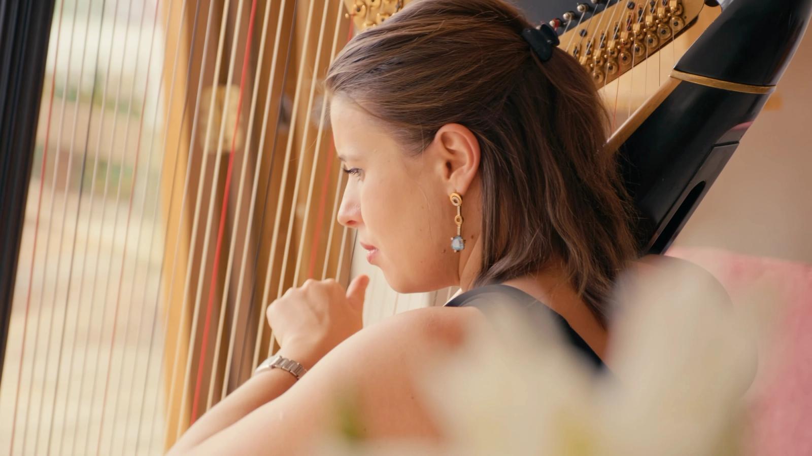 A woman with long brown hair, wearing a black top and gold earrings, sits on a pink sofa, playing a gold harp with black strings. She holds the harp w
