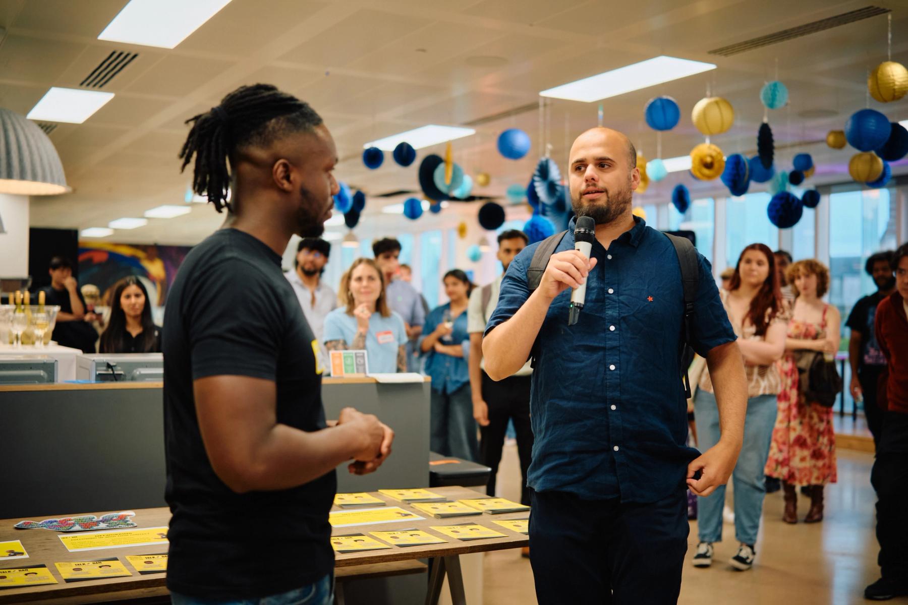Access VFX community event 2026, man in a blue shirt with a microphone talking beside a table of yellow flyers