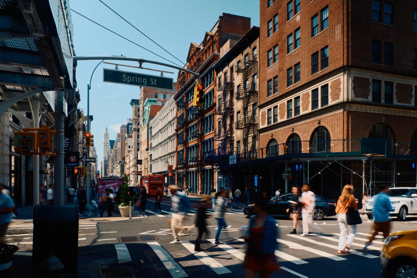 A bustling New York City street scene features brick buildings, a crosswalk, pedestrians, and cars.