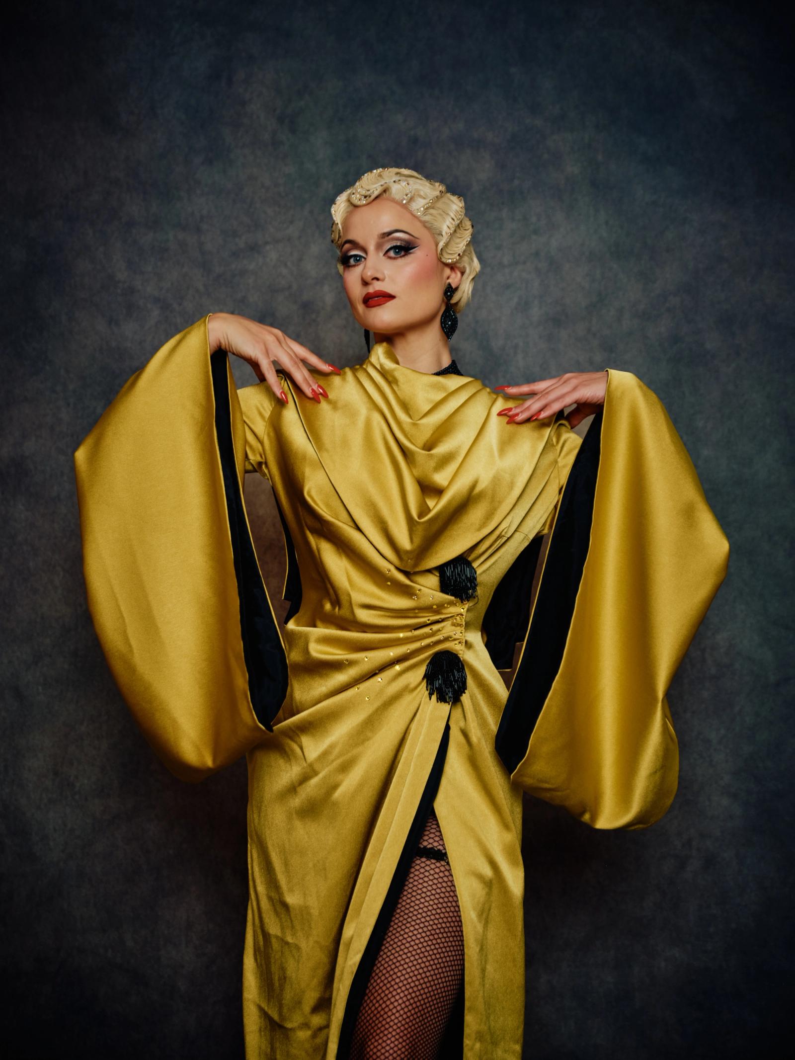 A woman in a vibrant yellow dress with black accents, adorned with a black belt and a black tassel, poses elegantly with her hands on her hips and her