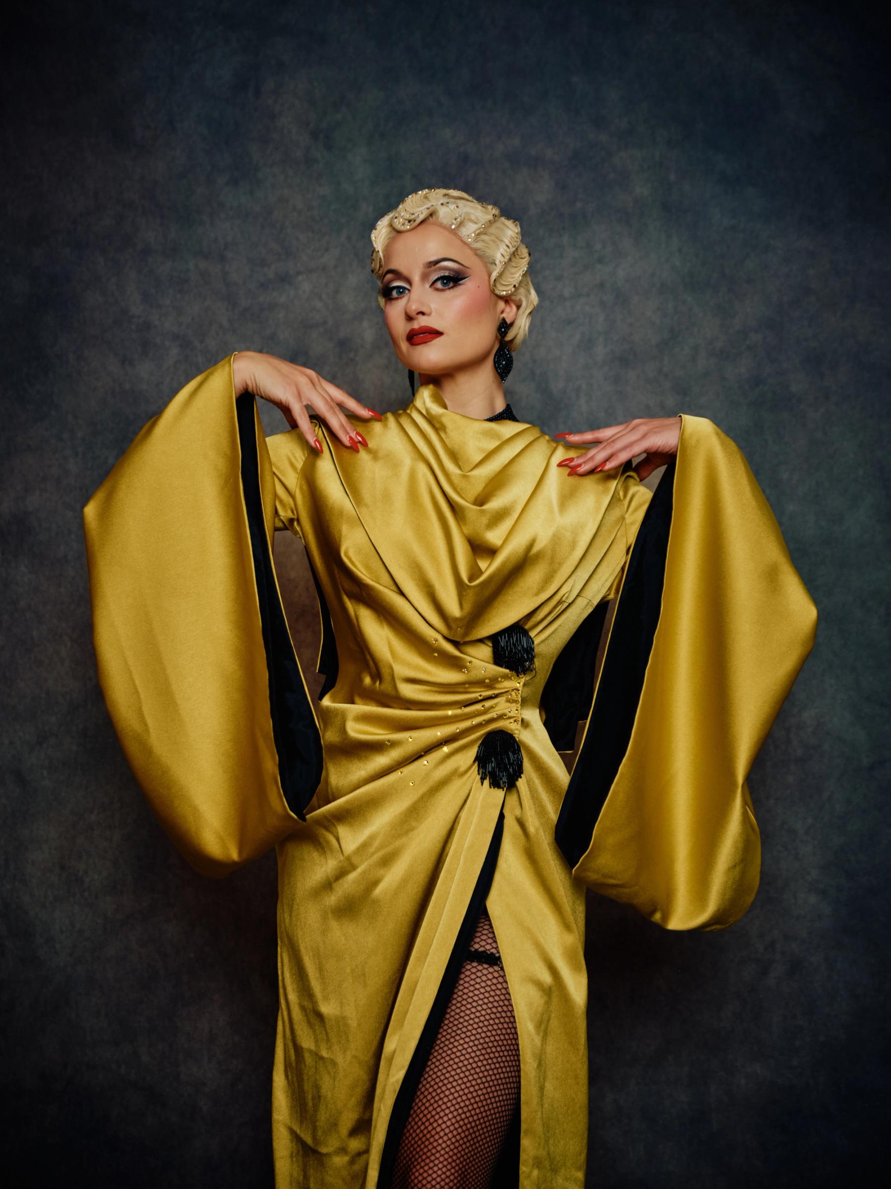 A woman in a vibrant yellow dress with black accents, adorned with a black belt and a black tassel, poses elegantly with her hands on her hips and her