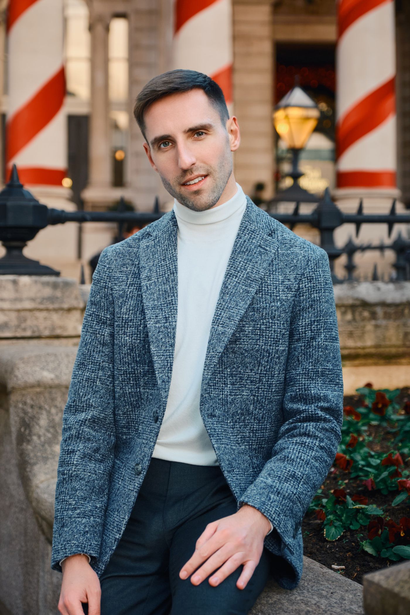 A man in a gray blazer and white turtleneck, wearing a white turtleneck and black pants, poses on a stone ledge in front of a building with red and wh