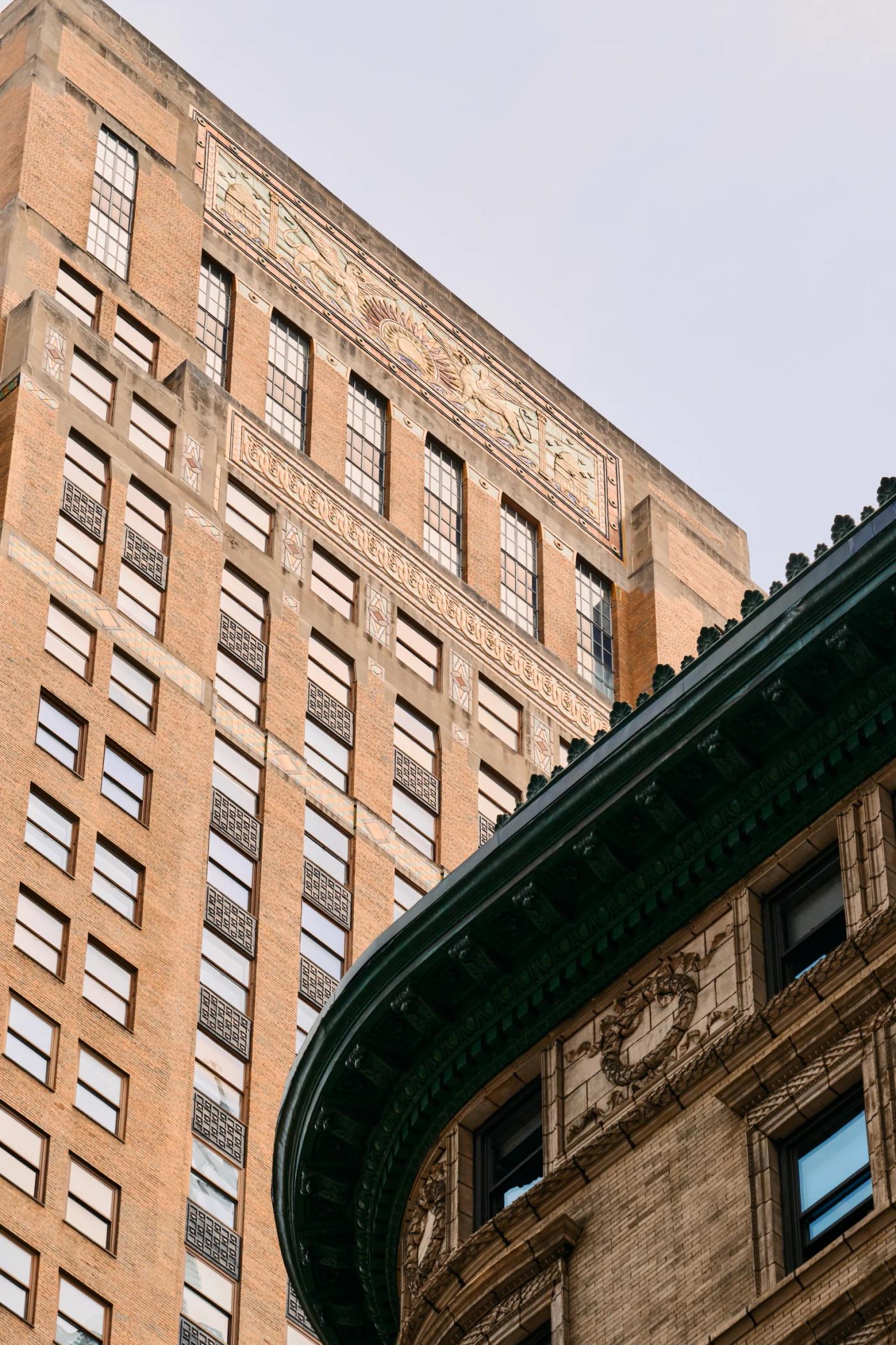 A tall brick building with a green roof and intricate carvings stands in a bustling city, its red bricks contrasting with the clear blue sky.