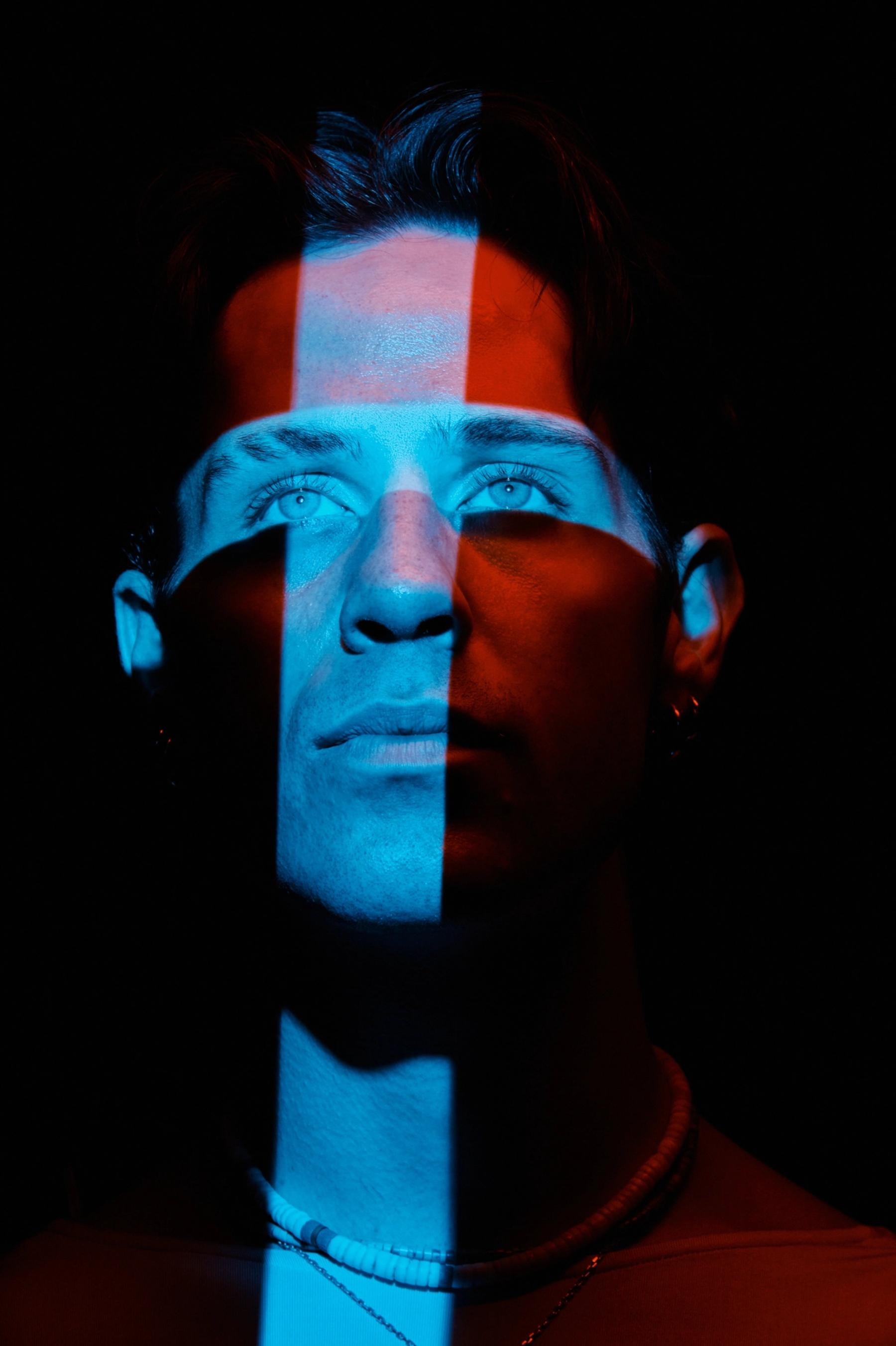 A man with a necklace and earrings, wearing a red shirt, is in a dark room with blue and red stripes on his face.