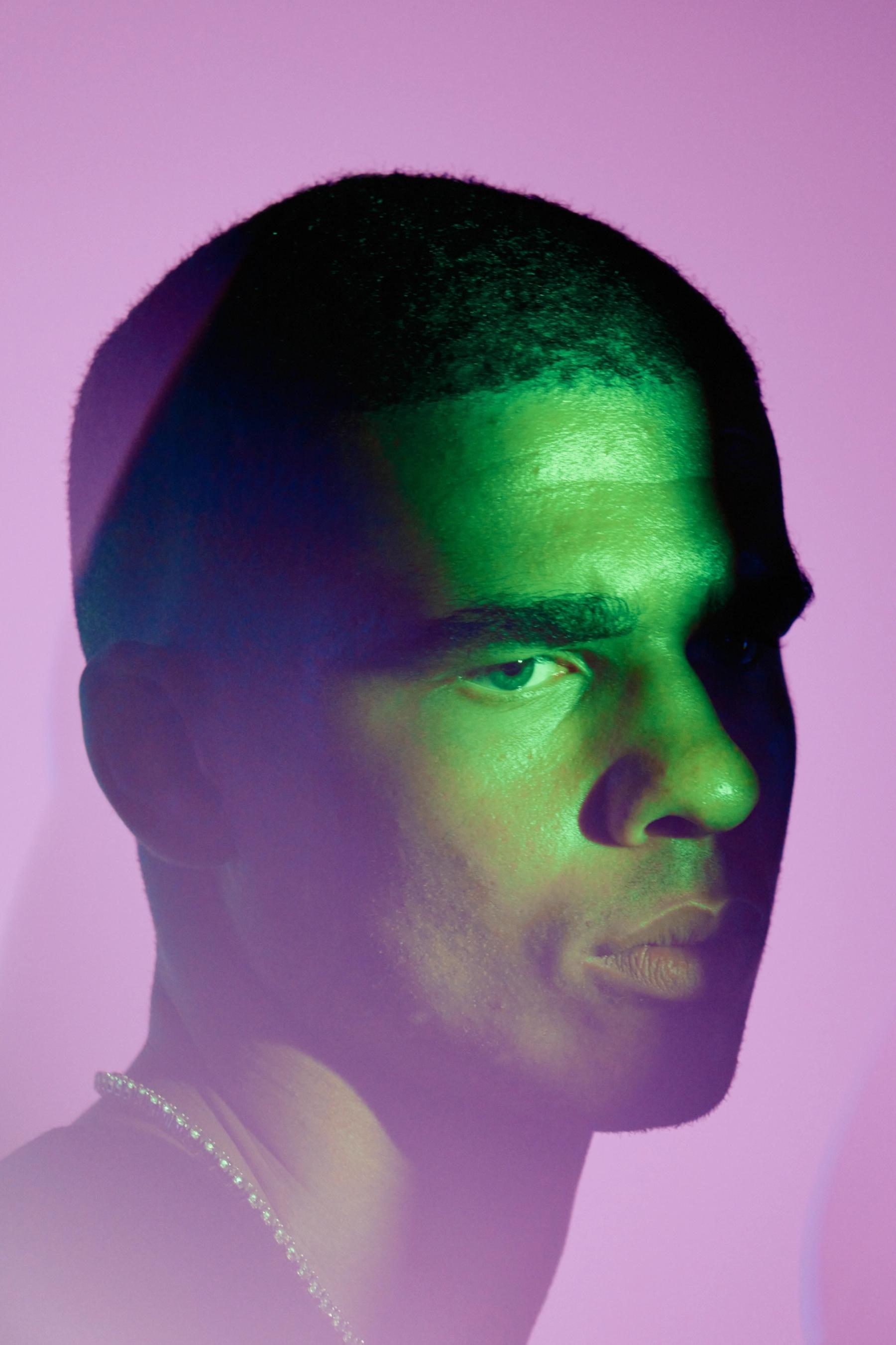 A man with a greenish hue, wearing a necklace, is looking at the camera with a serious expression.